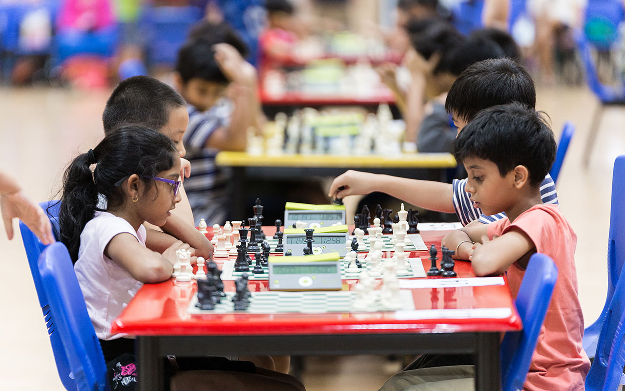 OFS Elementary School students sitting down in front of a chess set on a table during Elementary School chess tournament in auditorium