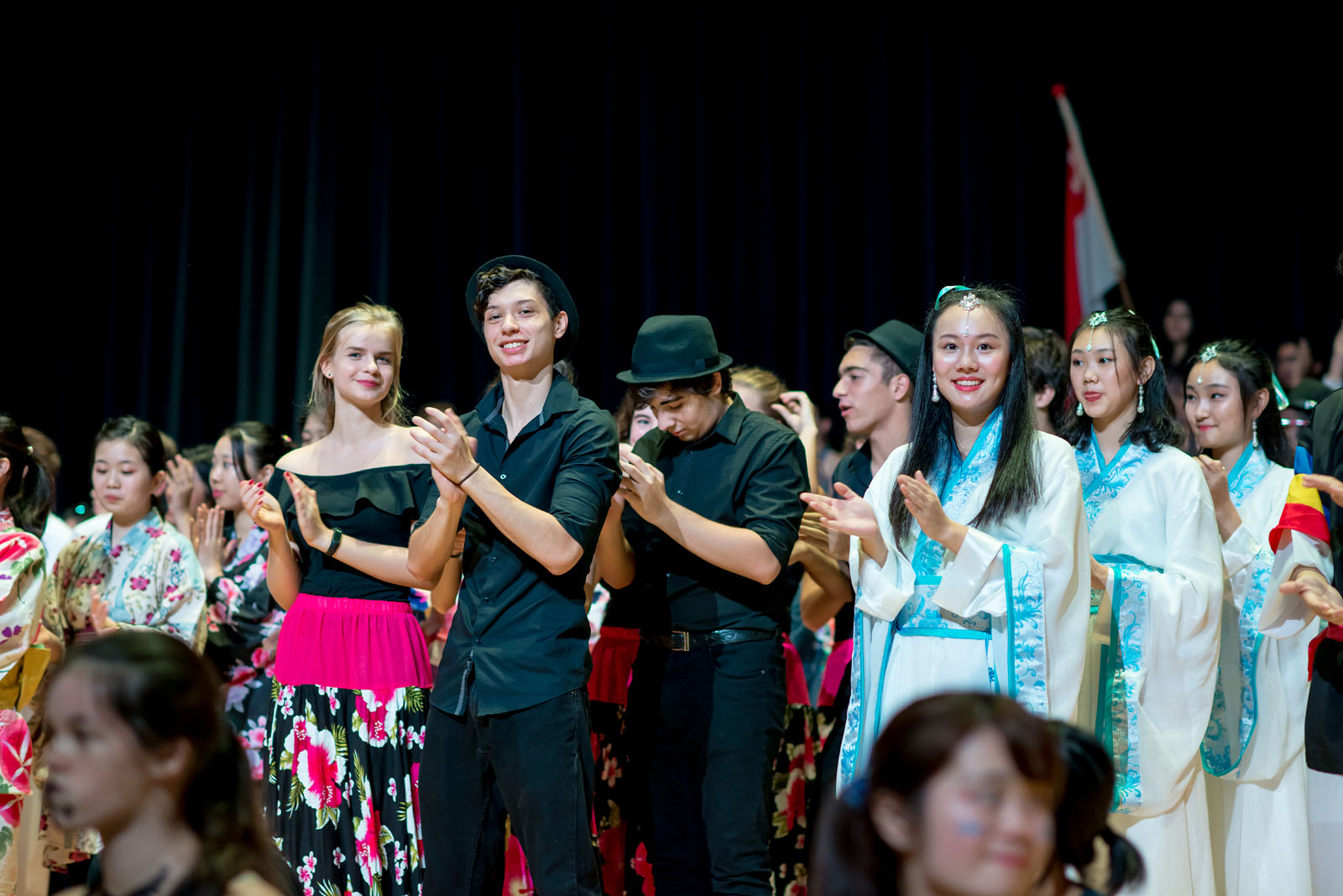 OFS High School students in their full performance costumes clapping hands on the auditorium stage after a memorable UN Night performance